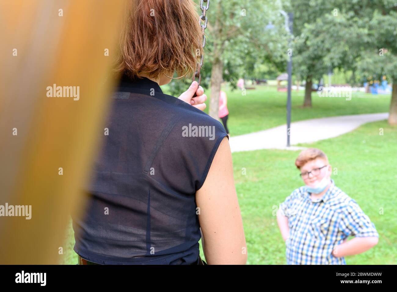 Junge, der Brille und Maske trägt, in einem karierten Hemd gekleidet, schaut auf ein Mädchen auf dem Spielplatz. Kind vor Mädchen in einem öffentlichen Park. Stockfoto