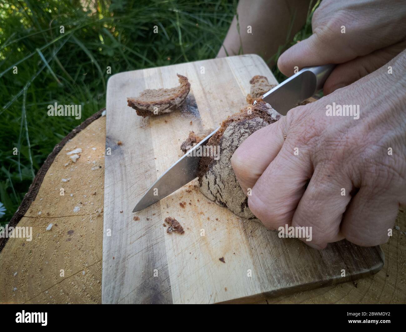 Detail der menschlichen Hände schneiden altes Brot mit Messer auf Holzbrett in Garten-Küche Stockfoto