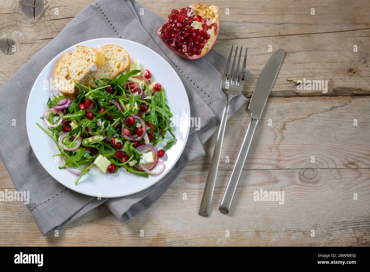 Rucola oder Rucola Salat mit Granatapfelkernen, roten Zwiebeln und Parmesan serviert auf einem weißen Teller mit Besteck auf einem rustikalen Holztisch, Kopierraum, hig Stockfoto