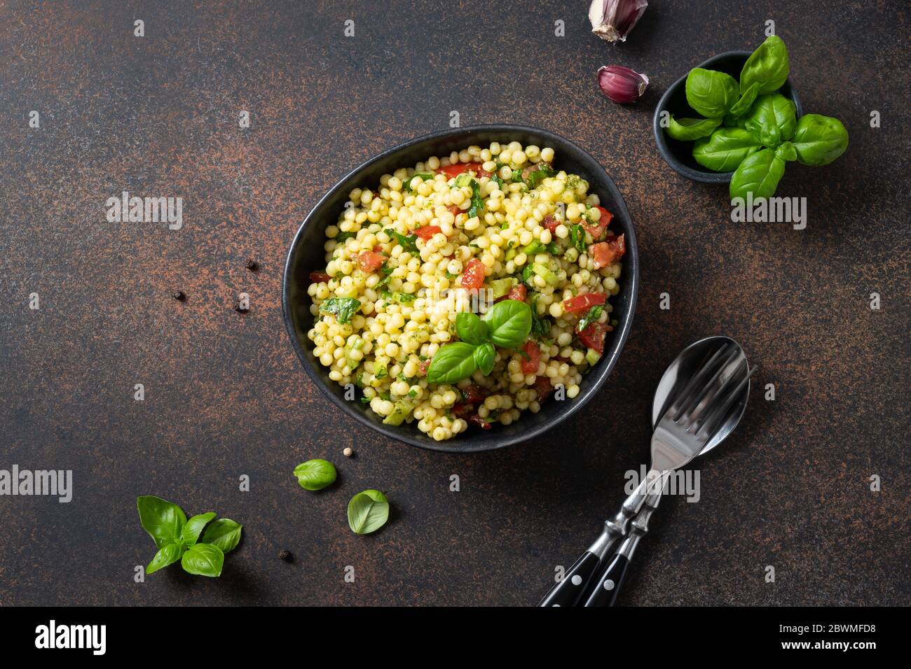 Gemüsesalat mit Pasta Ptitim oder Birdy, israelisch Couscous dekoriert Basilikumblätter auf dunkelbraunem Hintergrund. Draufsicht. Stockfoto