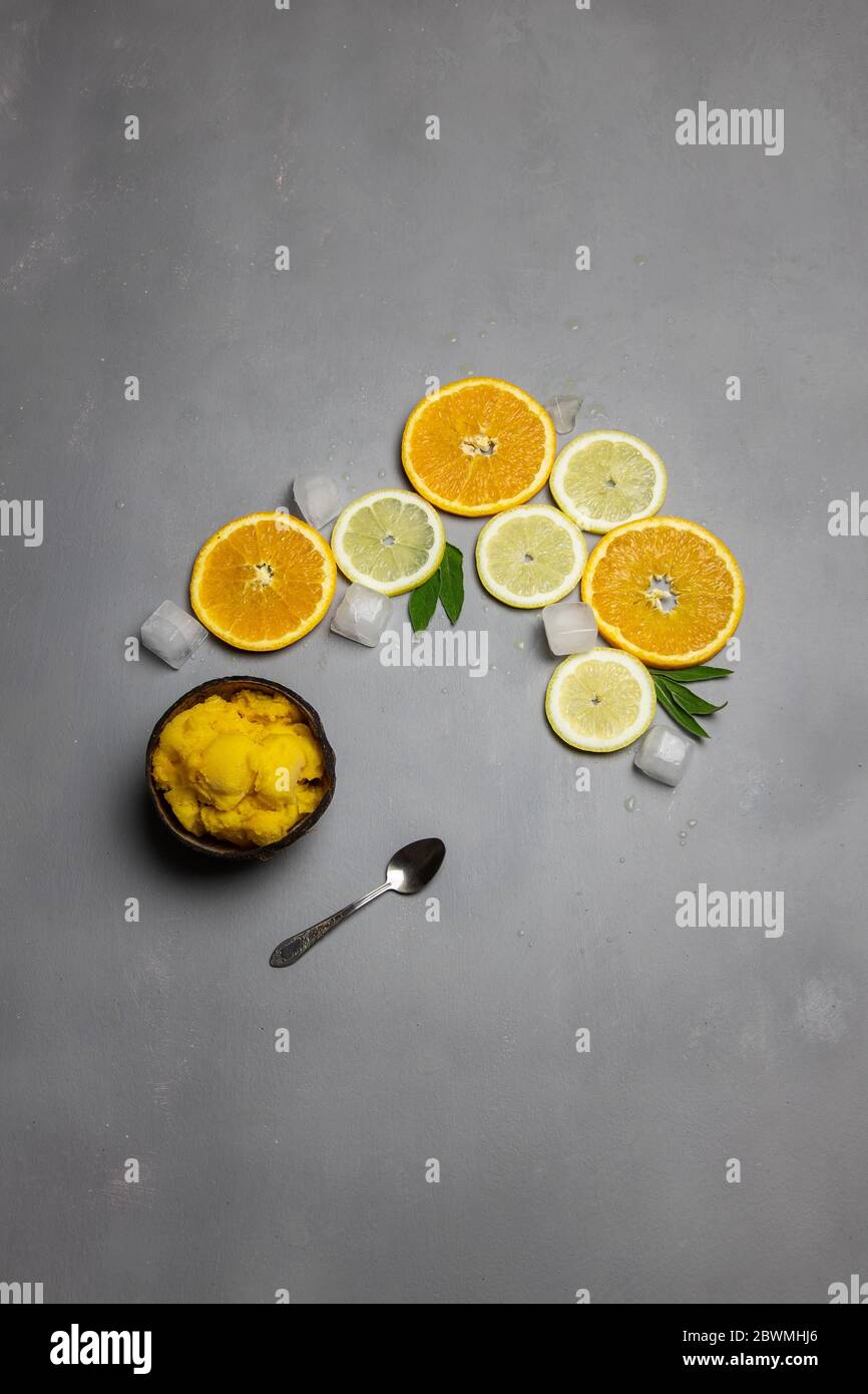 Kokosnussschale voll mit Zitrusfrüchten Sorbet Eis und kleinen metallischen Löffel, in Scheiben geschnittene Orangen und Zitronen auf der Seite mit Eiswürfeln auf grauem Beton Bac Stockfoto