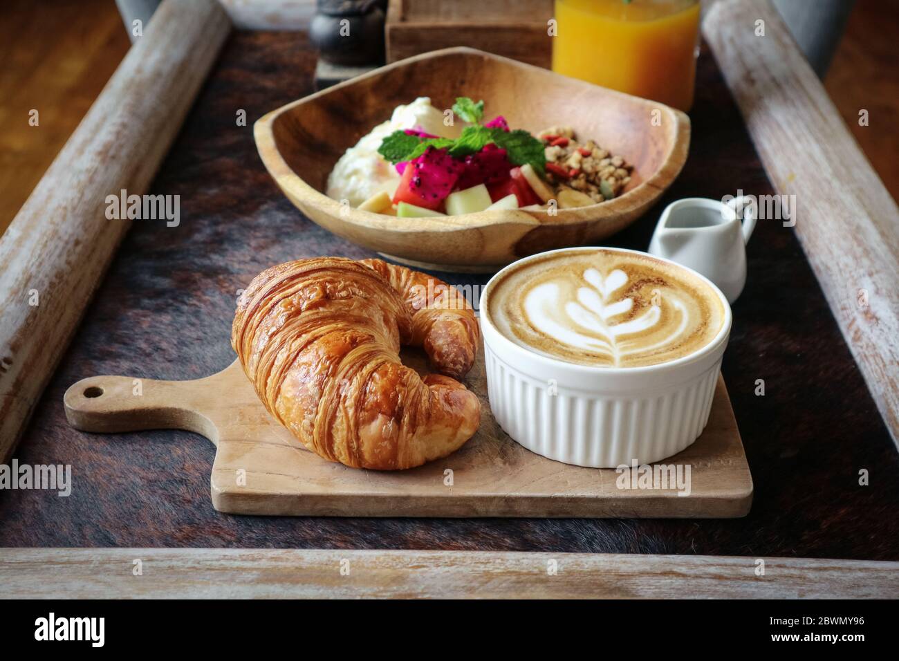 Eine Tasse Kaffee mit Latte Art auf dem Tisch, Buttercroissant und Müsli mit frischen tropischen Früchten und Joghurt auf dem Tisch im Café. Konzept der gesunden Brea Stockfoto