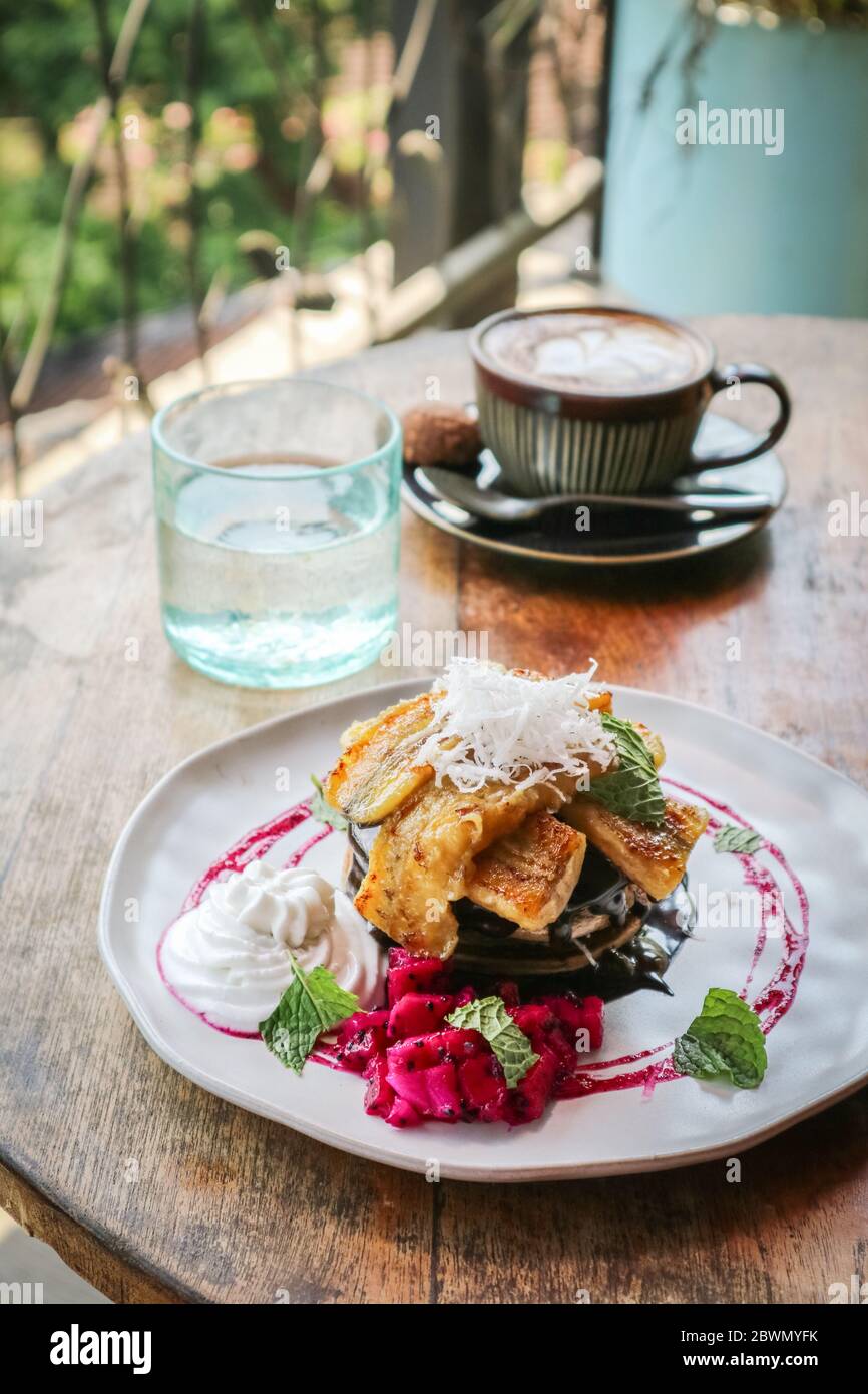 Stapel veganer Pfannkuchen mit Banane, Scheibe Drachenfrucht, Schokolade und Kokosnuss-Creme mit Kaffee und Wasser auf dem Tisch dekoriert Stockfoto