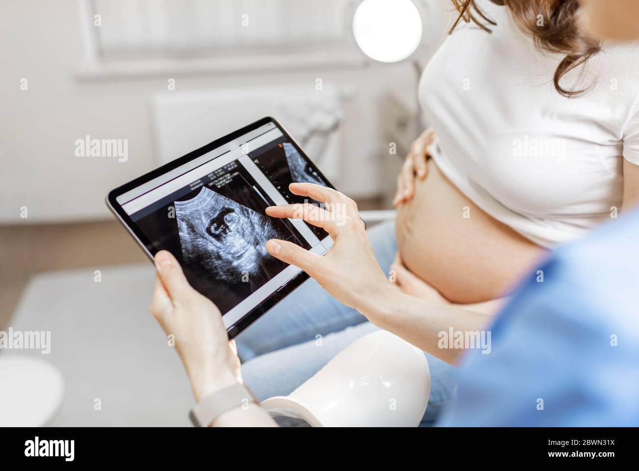 Arzt mit Ultraschall-Scan des ungeborenen Kindes auf einem digitalen Tablet während einer Untersuchung mit einer schwangeren Frau im Büro, beschnittene Ansicht ohne Gesichter Stockfoto