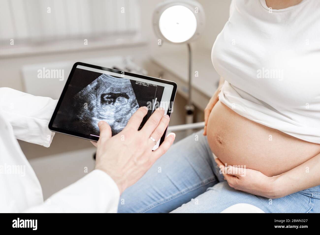 Arzt mit Ultraschall-Scan des ungeborenen Kindes auf einem digitalen Tablet während einer Untersuchung mit einer schwangeren Frau im Büro, beschnittene Ansicht ohne Gesichter Stockfoto