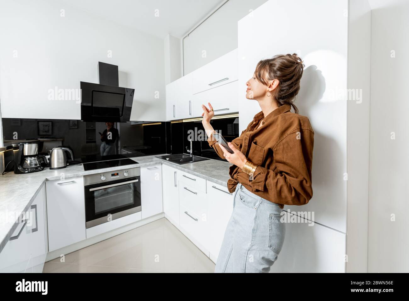 Junge Frau kontrolliert Küchengeräte mit Handy und Sprachbefehlen, breite Innenansicht. Smart Home-Konzept Stockfoto