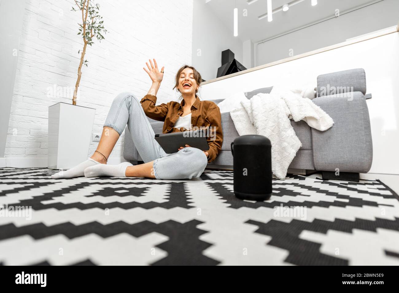Frau, die Sprachbefehle verwendet, um ein Smart-Home-Gerät zu steuern, das mit einem Smart-Lautsprecher und einem Tablet auf dem Boden im Wohnzimmer sitzt Stockfoto