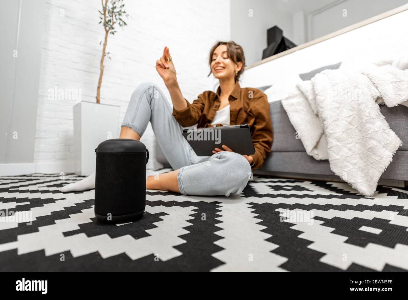 Frau, die Sprachbefehle verwendet, um ein Smart-Home-Gerät zu steuern, das mit einem Smart-Lautsprecher und einem Tablet auf dem Boden im Wohnzimmer sitzt Stockfoto