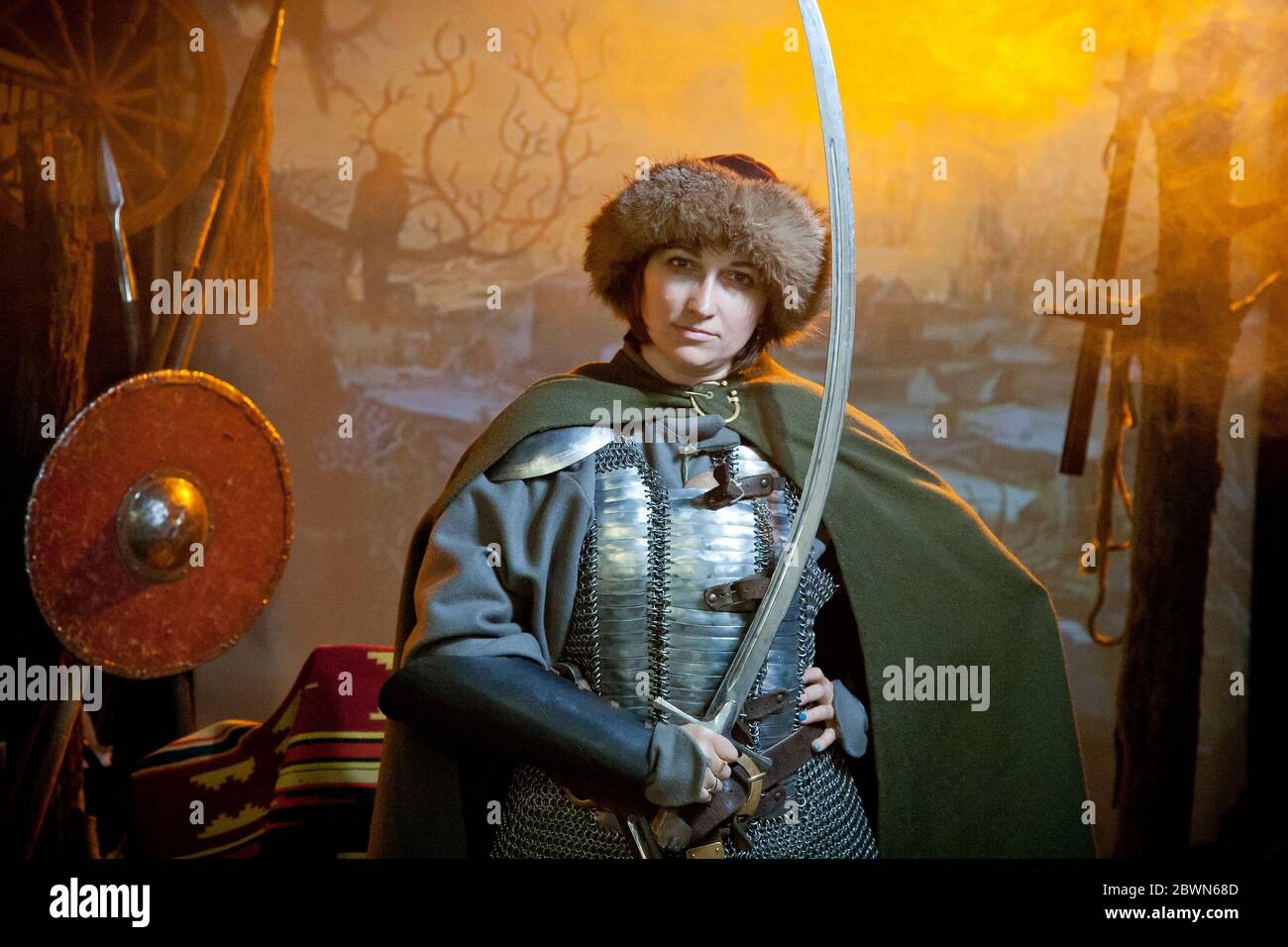 Eine Frau in Kettenpost, Mantel und Pelzhut gekleidet mit einem Säbel in den Händen vor dem Hintergrund von Waffen, einem Zweig mit Krähe und einem Schloss in Stockfoto