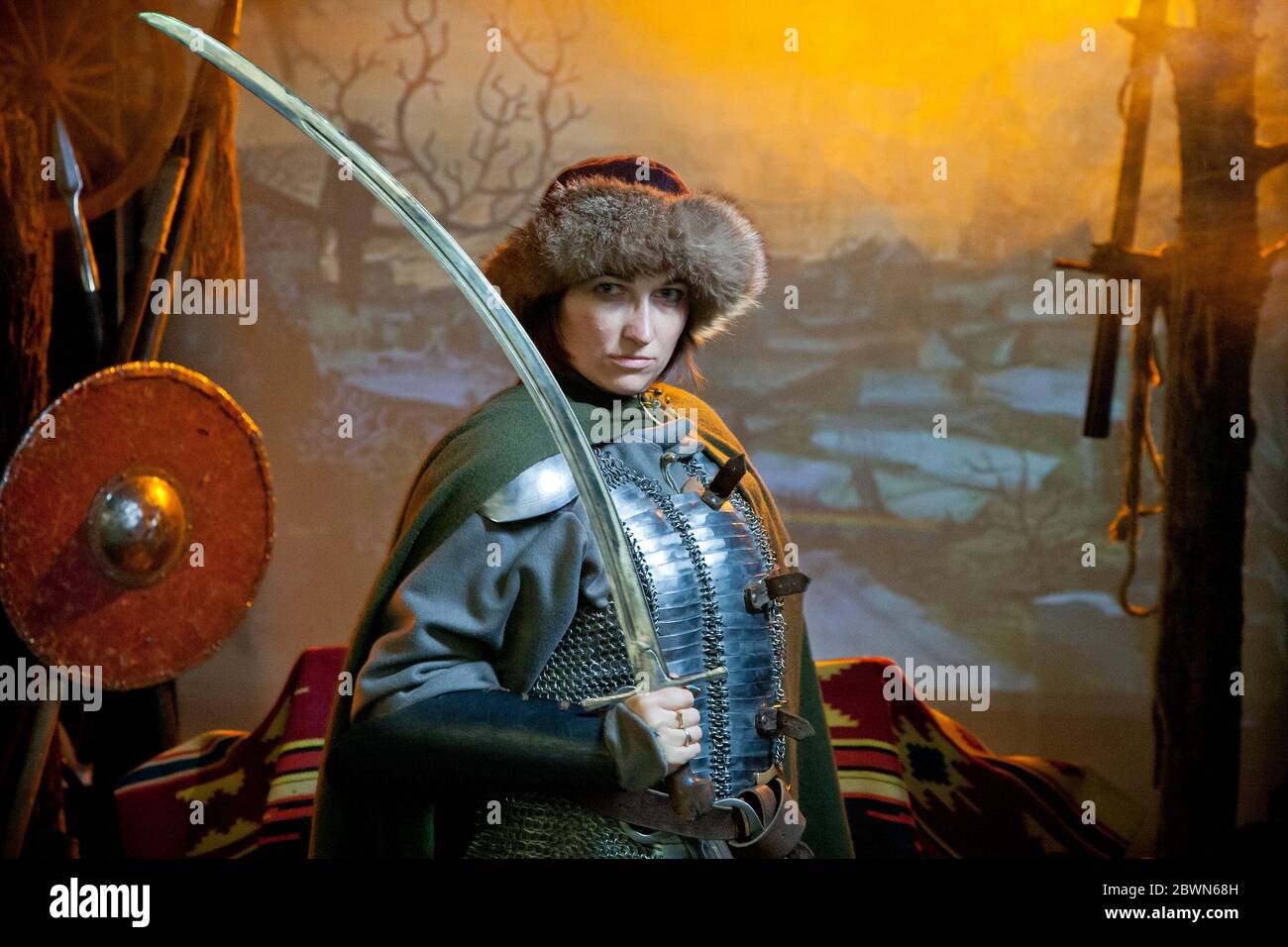 Eine Frau in Kettenpost, Mantel und Pelzhut gekleidet mit einem Säbel in den Händen vor dem Hintergrund von Waffen, einem Zweig mit Krähe und einem Schloss in Stockfoto