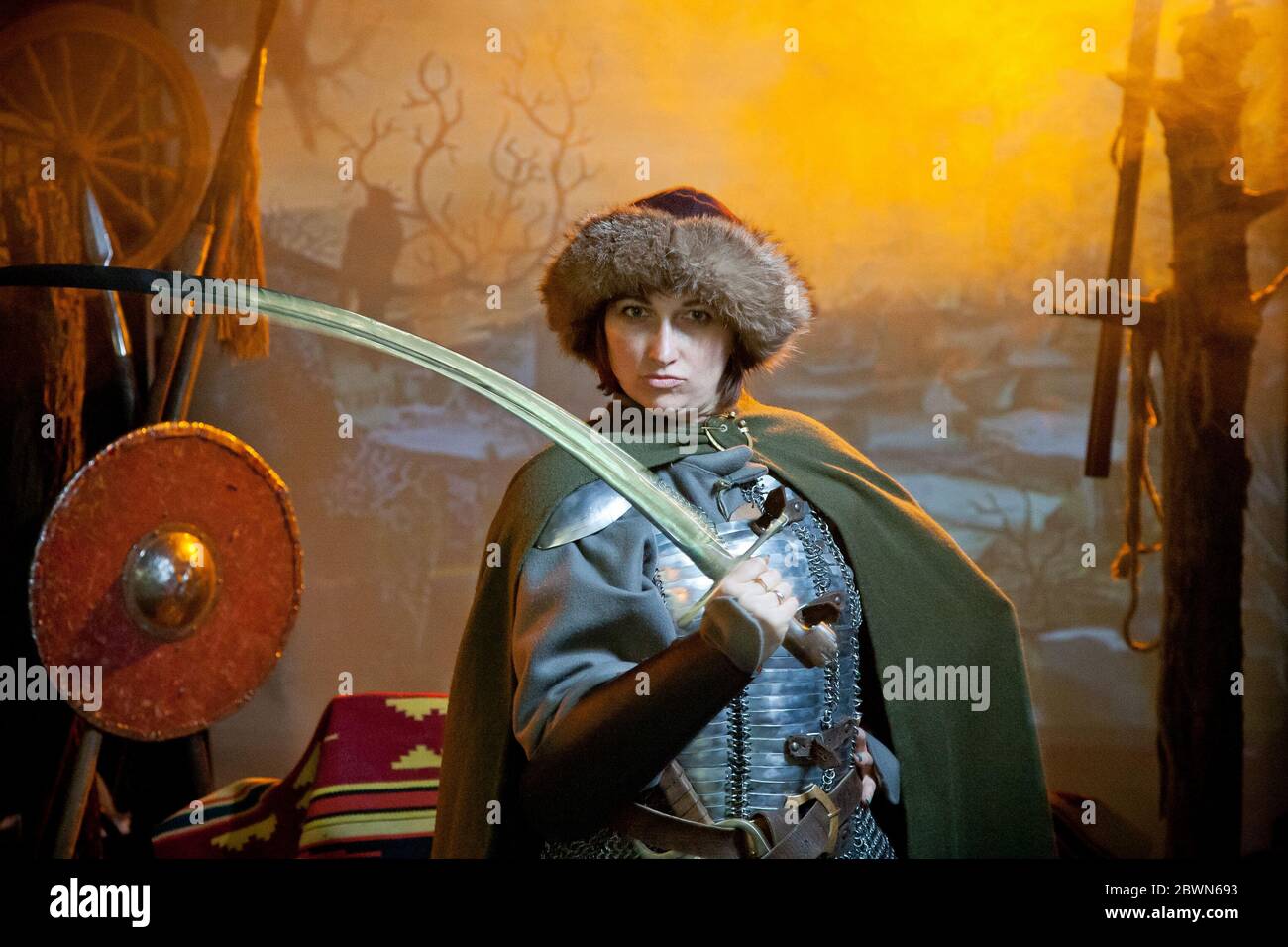 Eine Frau in Kettenpost, Mantel und Pelzhut gekleidet mit einem Säbel in den Händen vor dem Hintergrund von Waffen, einem Zweig mit Krähe und einem Schloss in Stockfoto