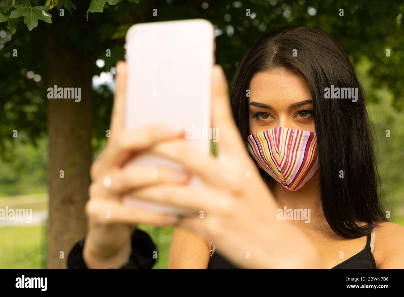 Junge Frau mit einer bunten modischen Gesichtsmaske, die ein Selfie auf ihrem Handy im Freien in einem Post Covid-19 Lifestyle-Konzept Stockfoto