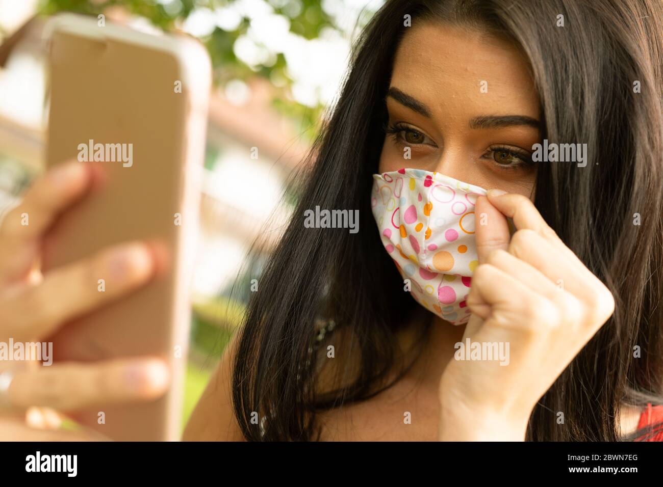 Junge Frau, die ihre bunte modische Gesichtsmaske mit ihrem Mobiltelefon im Freien in einem Garten oder Park während der Coronavirus Pandemie conceptu Stockfoto