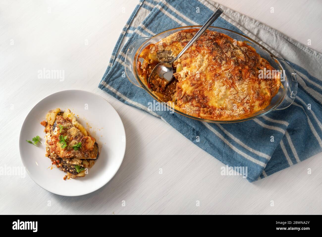 Moussaka, in einer Glasform auf einem blauen Küchentuch gebacken und auf einem weißen Teller serviert, traditionelle griechische Gericht mit Auberginen, Kartoffeln, Hackfleisch Stockfoto