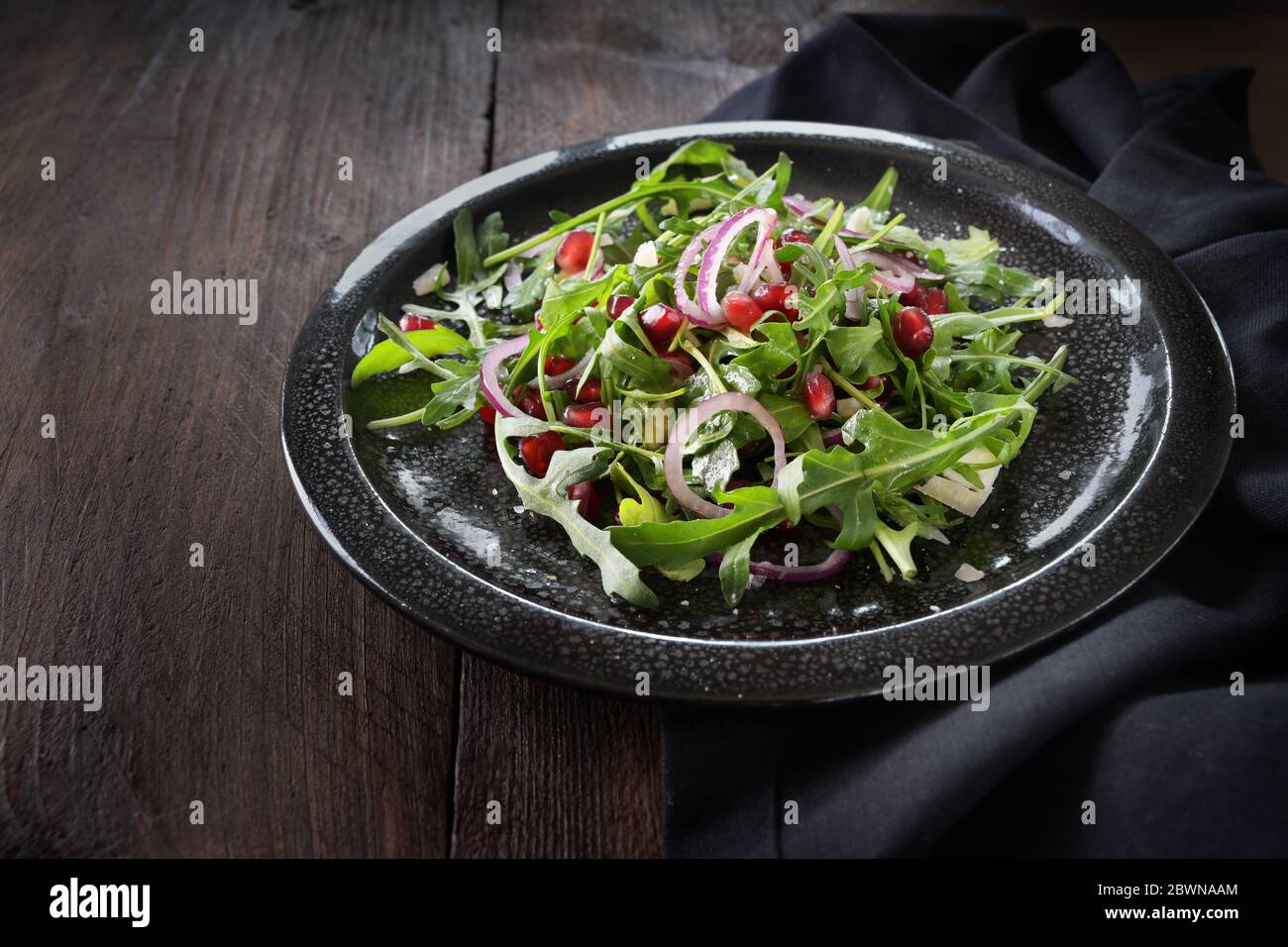 Frischer Rucola-Salat oder Rucola mit Granatapfelkernen, roten Zwiebeln und Feta-Käse auf einem schwarzen Gefleckteller auf einem dunklen Holztisch, Kopierraum, ausgewählt Stockfoto