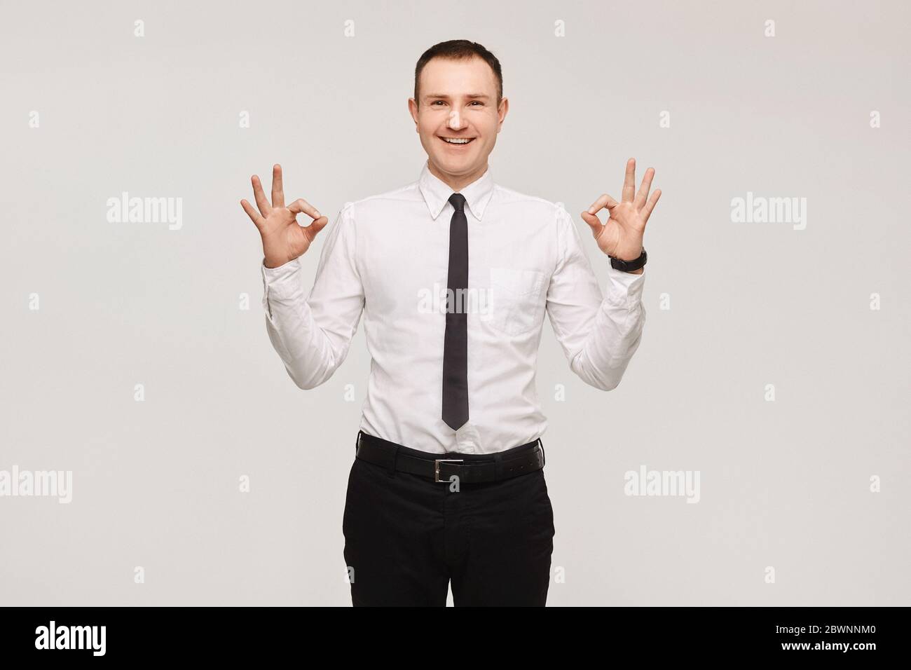 Gutaussehender lächelnder Geschäftsmann in einem weißen Hemd und schwarzer Krawatte zeigt ein OK-Zeichen mit Fingern über weißem Hintergrund, isoliert. Werben Sie für ein Modell Stockfoto