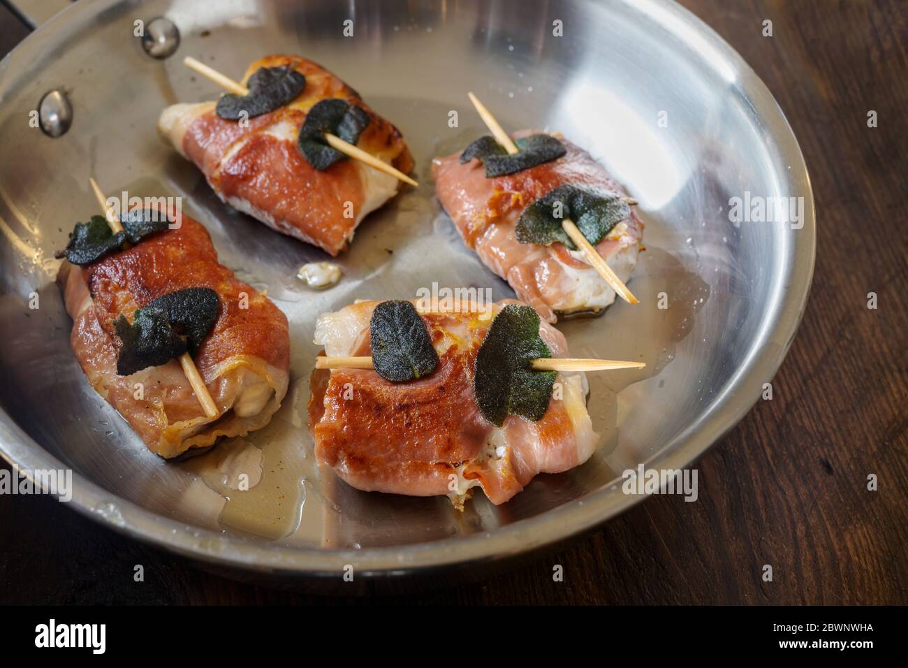 Gebratenes Hähnchen Saltimbocca mit Schinken und Salbeiblättern in einer Edelstahlpfanne auf rustikalem Holz, ausgewählter Fokus, schmale Schärfentiefe Stockfoto