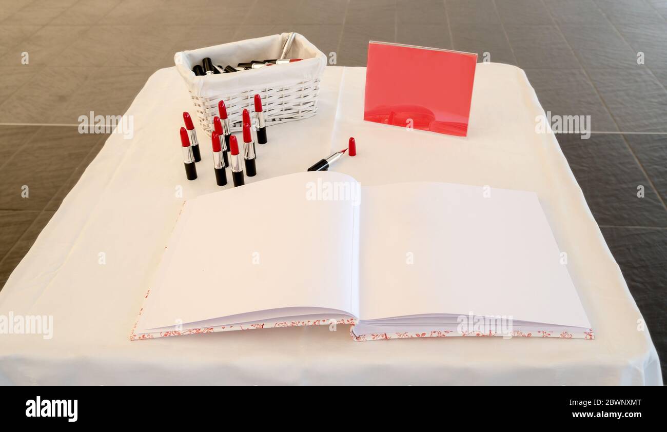 Leere Seiten eines Signaturbuchs auf einem Tisch neben Lippenstift-förmigen Stiften und einem roten Plakat mit Kopierfläche. Stockfoto