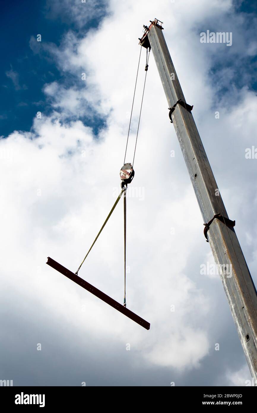 Baukran mit einem Haken und einem Metallprofil, das an Seilen hängt, die hochheben, niedrige Winkelansicht mit wolkenverhangen, sonnigen blauen Himmel Stockfoto