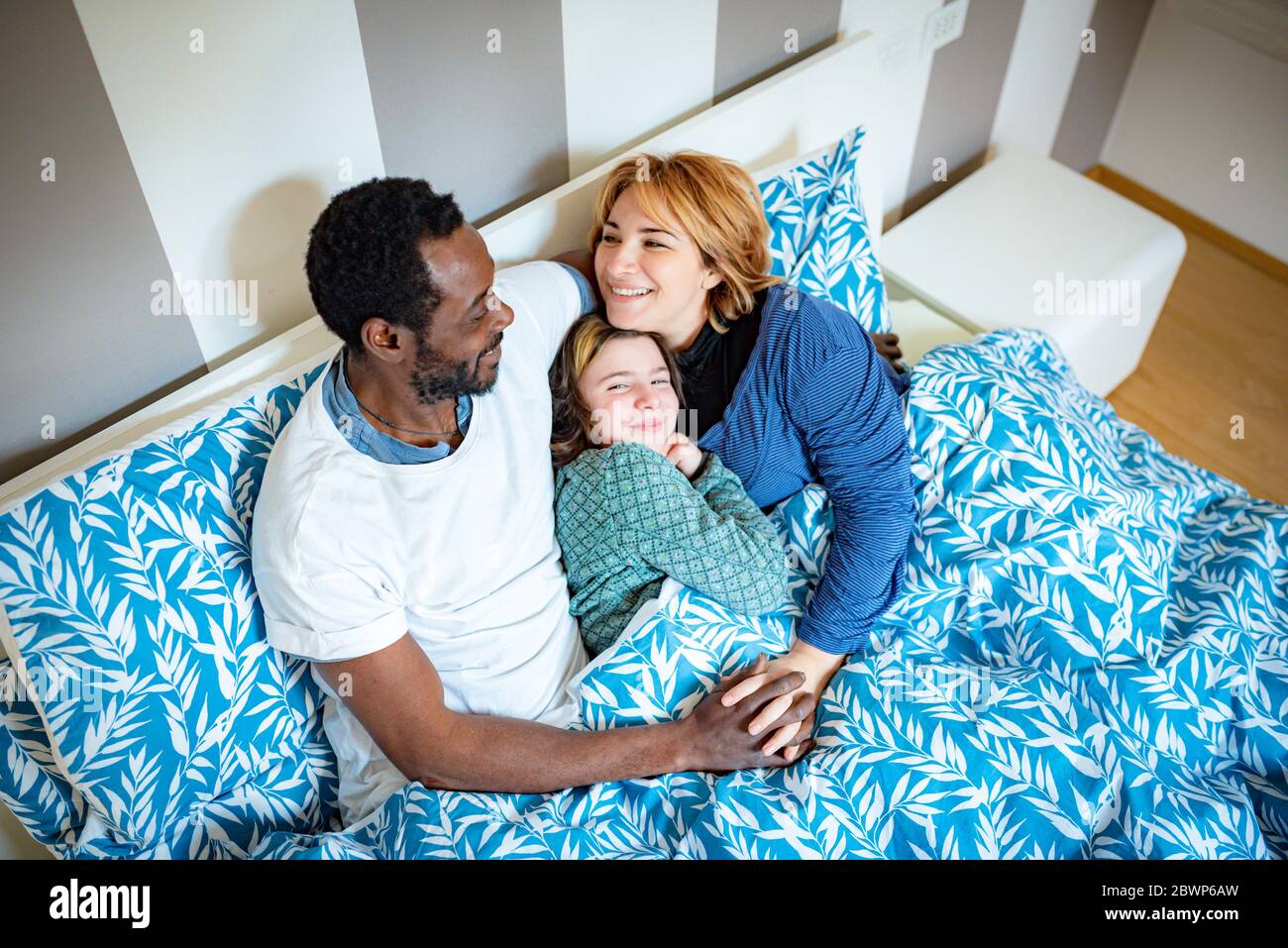 Glücklich multi ethnischen Familie Umarmungen im Bett Sobald sie aufwachen, kaukasische Mutter und afrikanischen Vater umarmen ihre Tochter zärtlich, kuscheln im Bett in den mornin Stockfoto