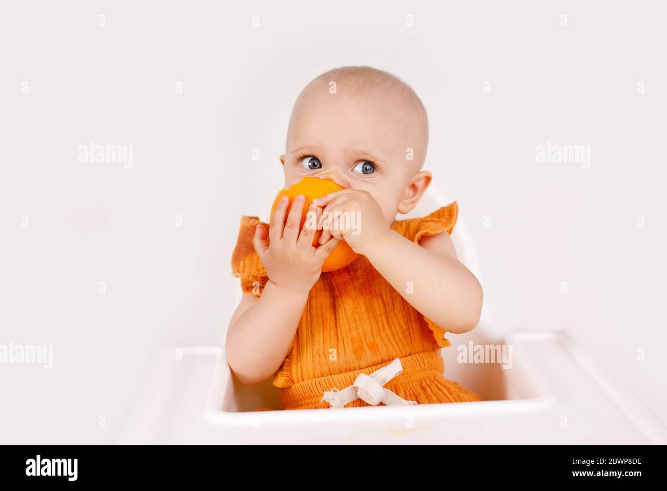 Glückliches Baby Mädchen, das im Hochstuhl sitzt und Orangen in einer weißen Küche isst. Gesunde Ernährung für Kinder. Bio-Früchte als erste feste Nahrung für Säuglinge Stockfoto