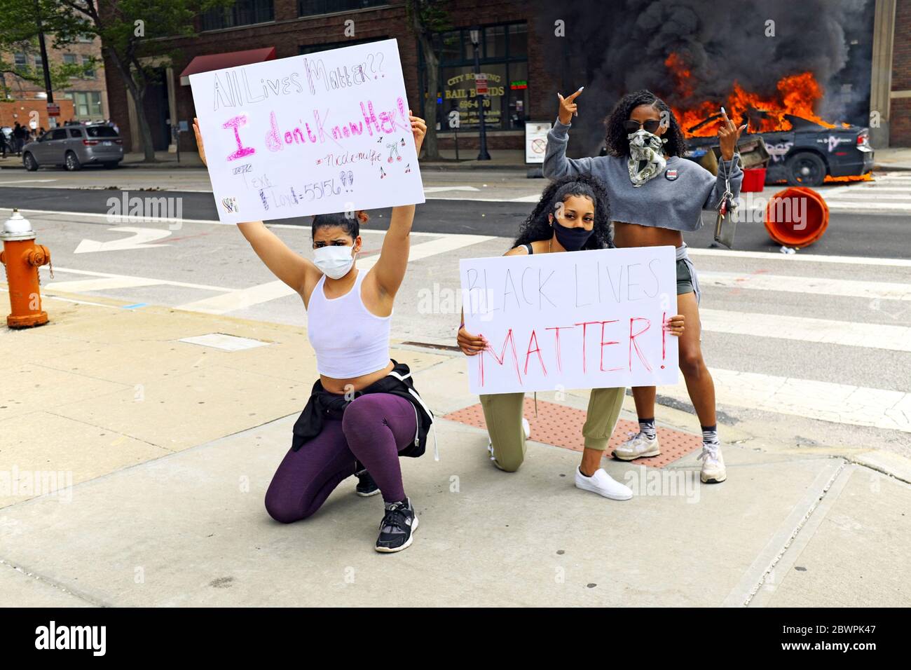 Drei Frauen posieren mit Schildern vor zwei brennenden Polizeiautos in der Innenstadt von Cleveland, Ohio, USA während eines Protestes zu Black Lives Matter am 30. Mai 2020. Die drei waren unter Tausenden, die in Cleveland auf die Straßen gingen, um gegen die Polizeipolitik, die Praktiken und die Ermordung schwarzer Menschen zu protestieren. Stockfoto
