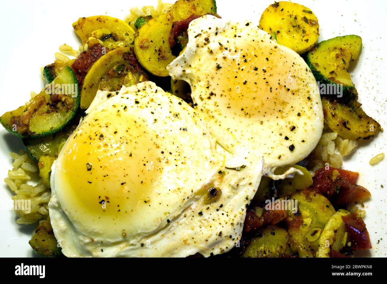 Zwei pochierte Eier liegen auf einem Bett aus gebratenen Zucchini, Sommer-Kürbis, Tomaten und braunem Reis. Stockfoto