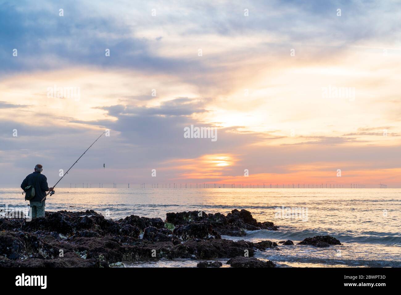 Sonnenaufgang über dem Meer bei Broadstairs an der Küste bei Ebbe. Einstehender Mann auf Felsen mit Angelrute im Vordergrund, der Sonnenaufgang halb von Wolken am Horizont verdeckt. Windparkturbinen am Horizont. Stockfoto