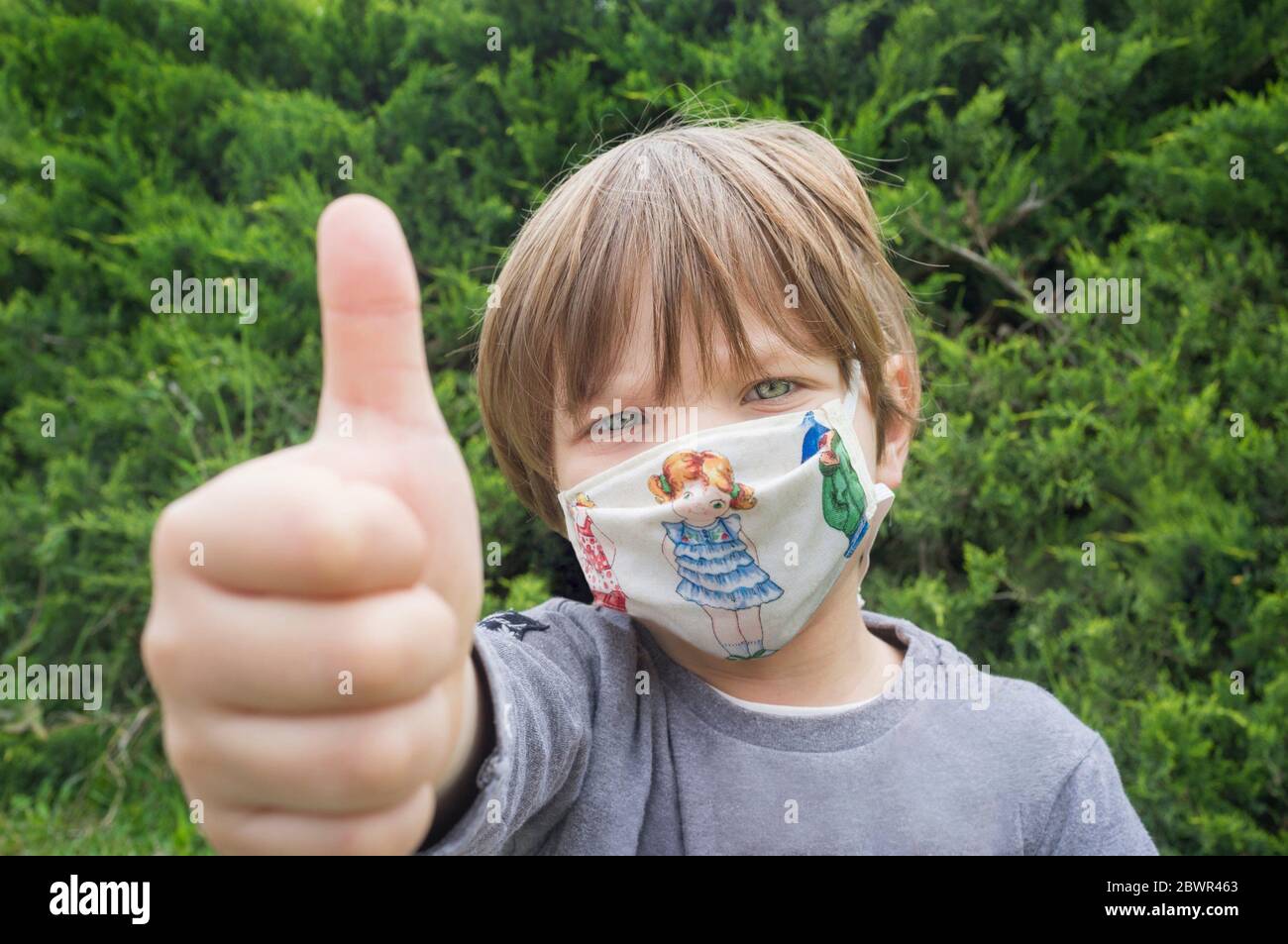 4 Jahre kleiner Junge mit Gesichtsmaske Print mit Kindermotiven. Er ist mit den Daumen nach oben Geste. Stockfoto