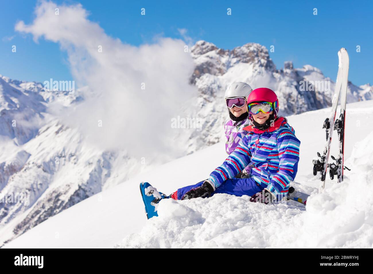 Zwei glückliche Mädchen sitzen in den Berg mit Ski über hohe Gipfel und Wolken im Schnee Blick auf Kamera lächelnd Stockfoto