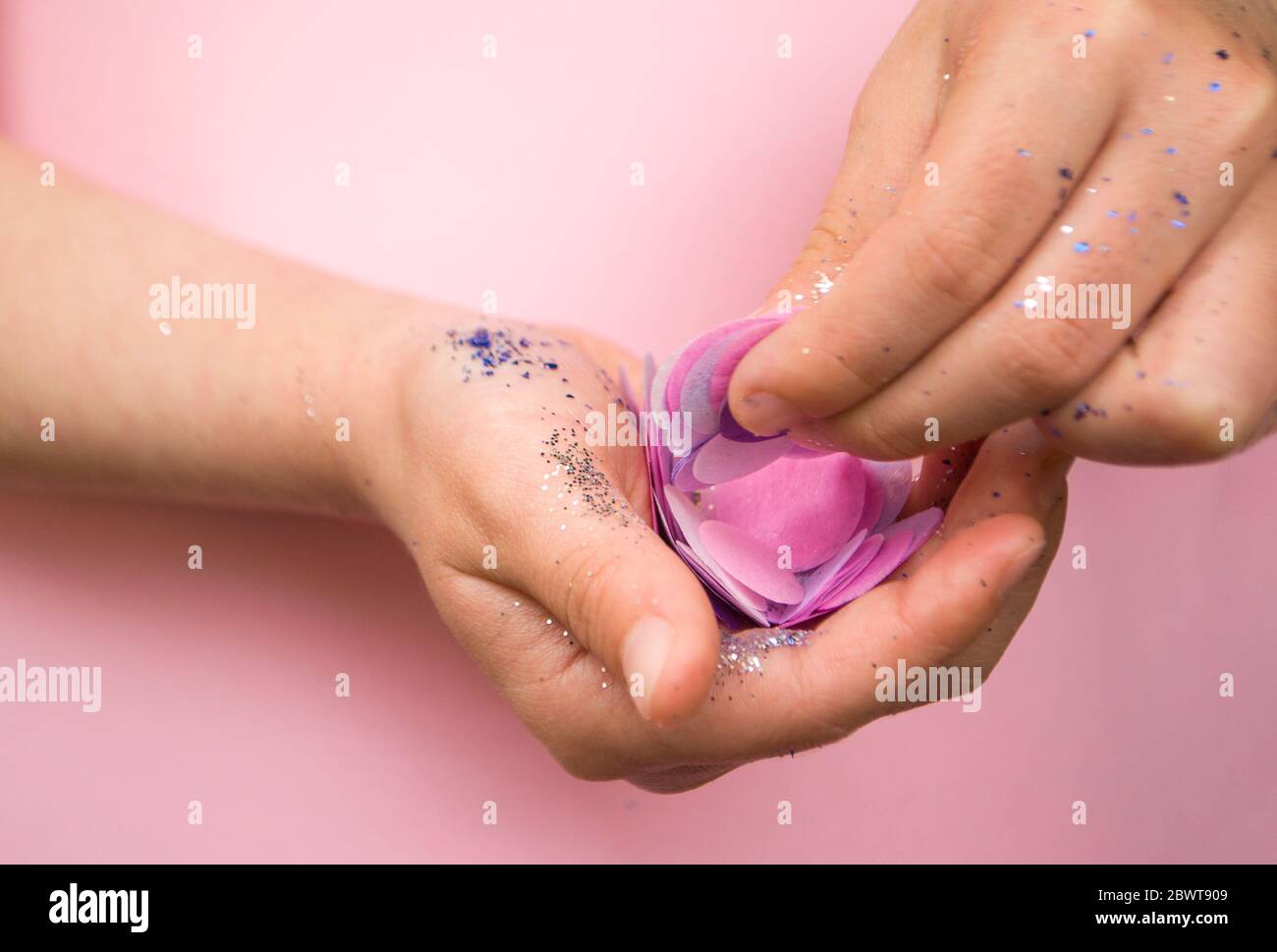 Kinderhände mit blauen und silbernen Glitzer und Konfetti auf pastellrosa Hintergrund. Stockfoto