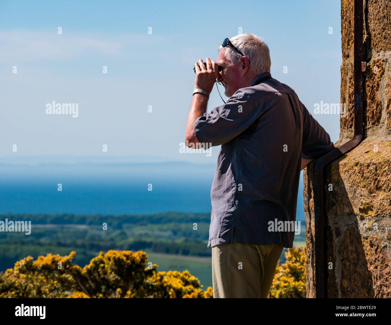 Senior Mann schaut durch Fernglas an sonnigen Tag in Sicht, East Lothian, Schottland, Großbritannien Stockfoto