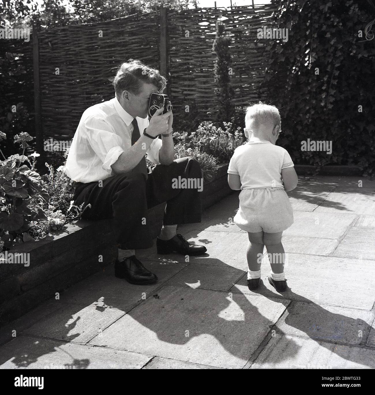 1960er Jahre, historisch, draußen auf einer Terrasse, ein Vater sitzt auf dem Rand eines Blumenbeds mit einer leichten Cine-Kamera, um seinen kleinen Sohn, England, Großbritannien zu Filmen. Kleine, tragbare 8-mm-Cine-Filmkameras waren in dieser Zeit beliebt für die Aufnahme bewegter Bilder des Alltags der Menschen und für Amateurfilme, die als "Heimfilme" bekannt sind. Stockfoto