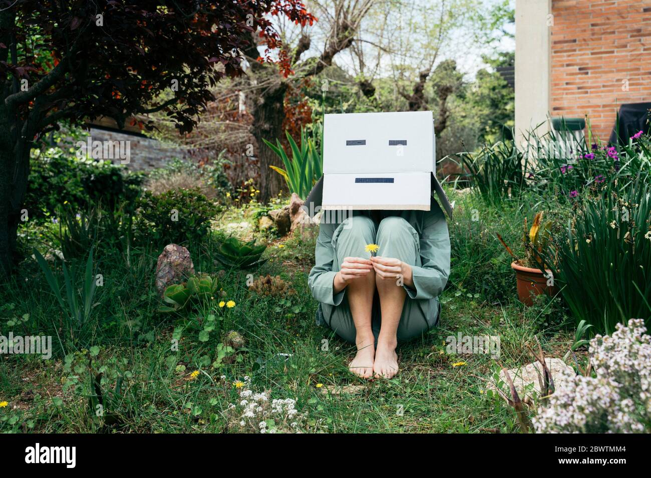 Junge Frau mit Pappkarton auf dem Kopf, barfuß im Garten sitzend Stockfoto