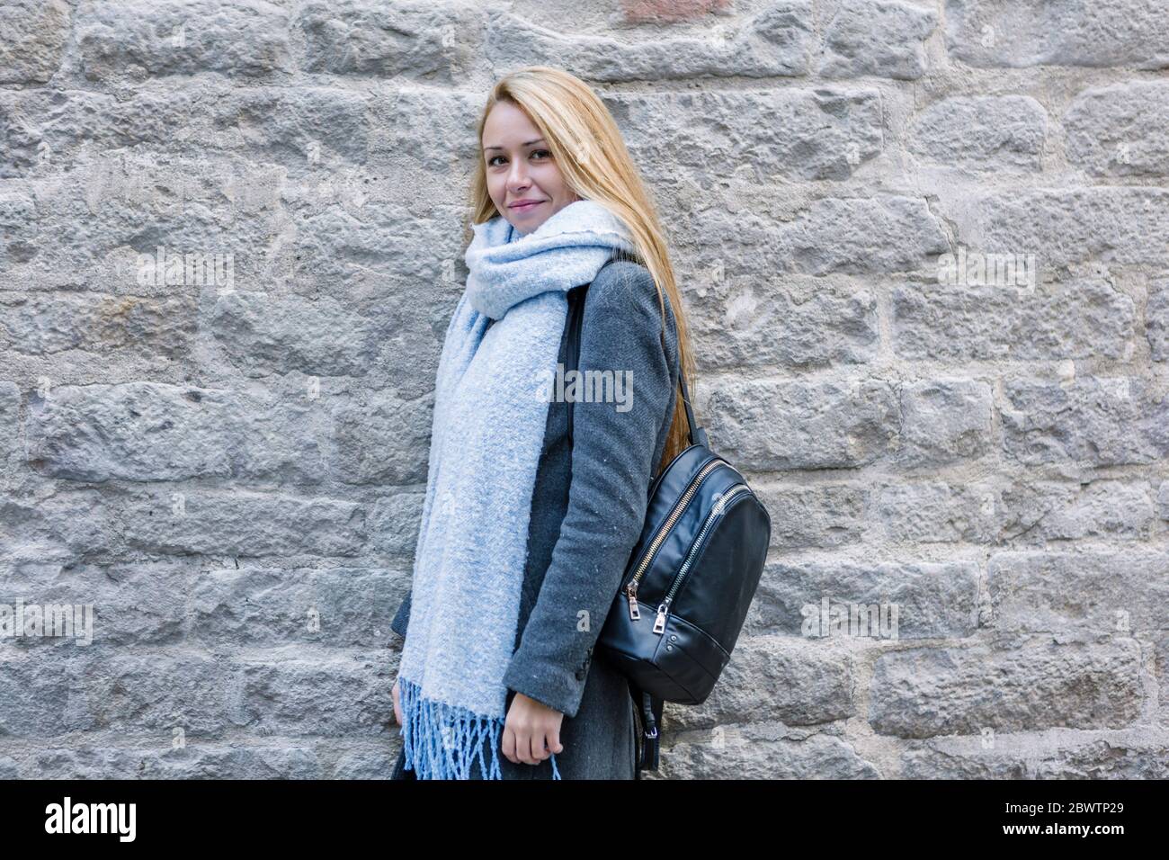 Portrait einer jungen Frau mit hellblauem Schal und Rucksack vor der Steinwand Stockfoto