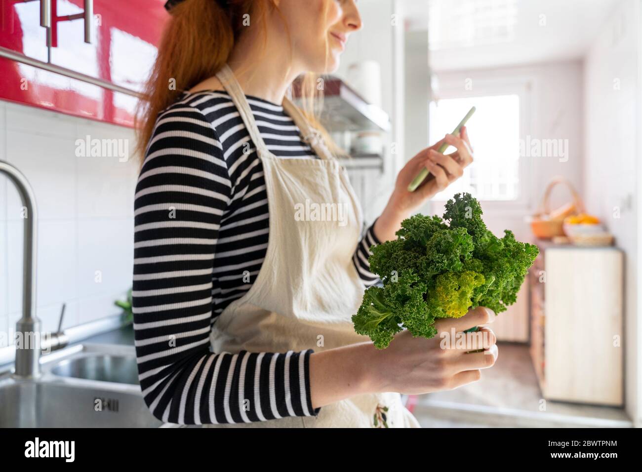 Junge Frau in der Küche stehend, halten Cale, mit dem Smartphone Stockfoto