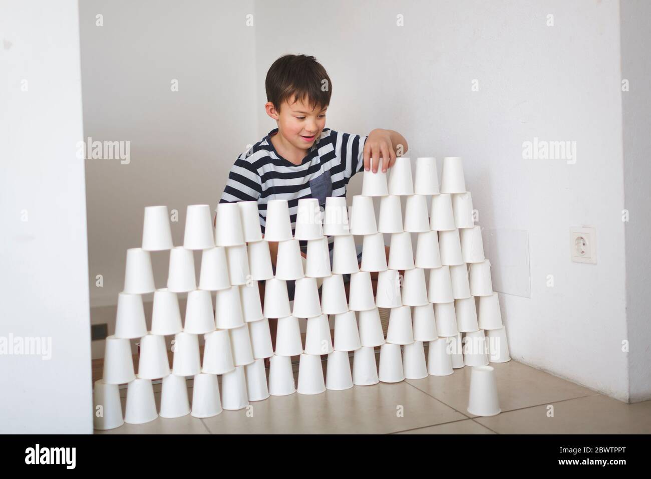 Lächelnder Junge, der zu Hause eine Wand aus weißen Pappbechern baute Stockfoto
