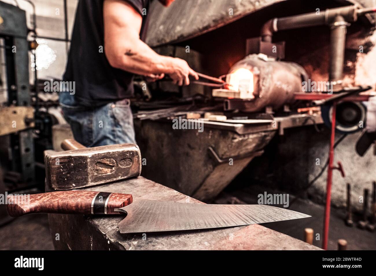 Messermacher arbeitet am Schmelzofen, fertiges Messer auf Amboss im Vordergrund Stockfoto