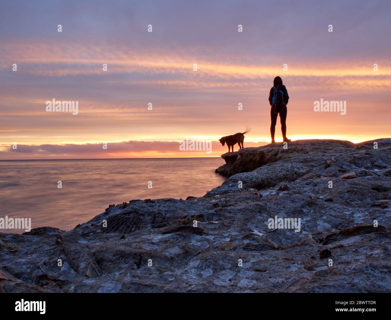 Rückansicht einer Frau, die mit Hund auf Felsformation am Meer während Sonnenuntergang steht, Puerto Piramides, Argentinien Stockfoto