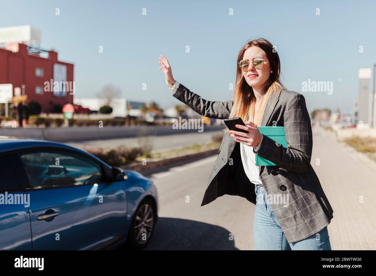 Junge Geschäftsfrau mit digitalem Tablet und Smartphone steht am Straßenrand und hagelt Taxi Stockfoto