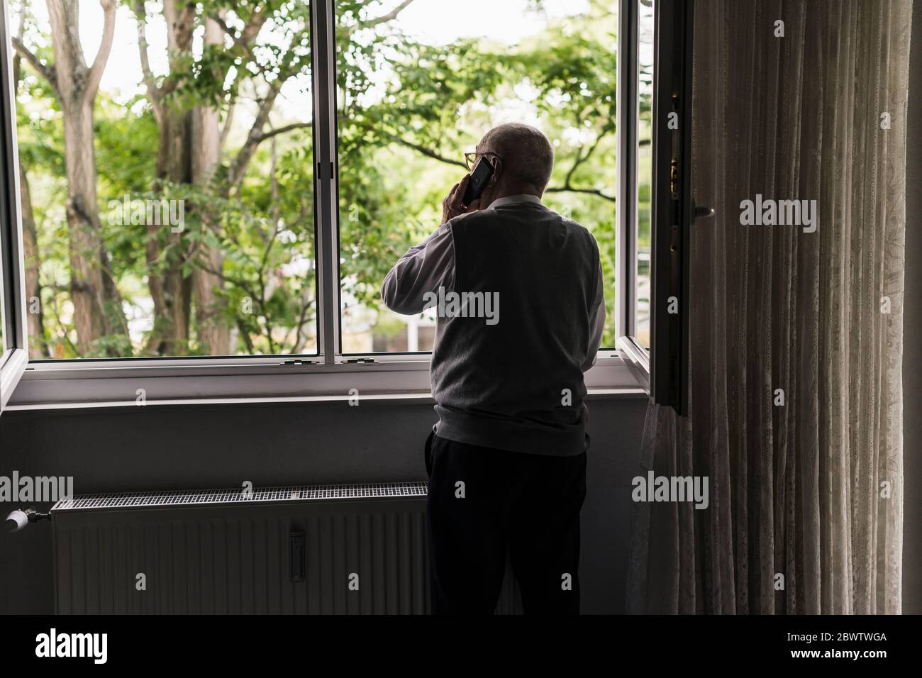 Rückansicht eines älteren Mannes am Telefon, der am offenen Fenster stand Stockfoto