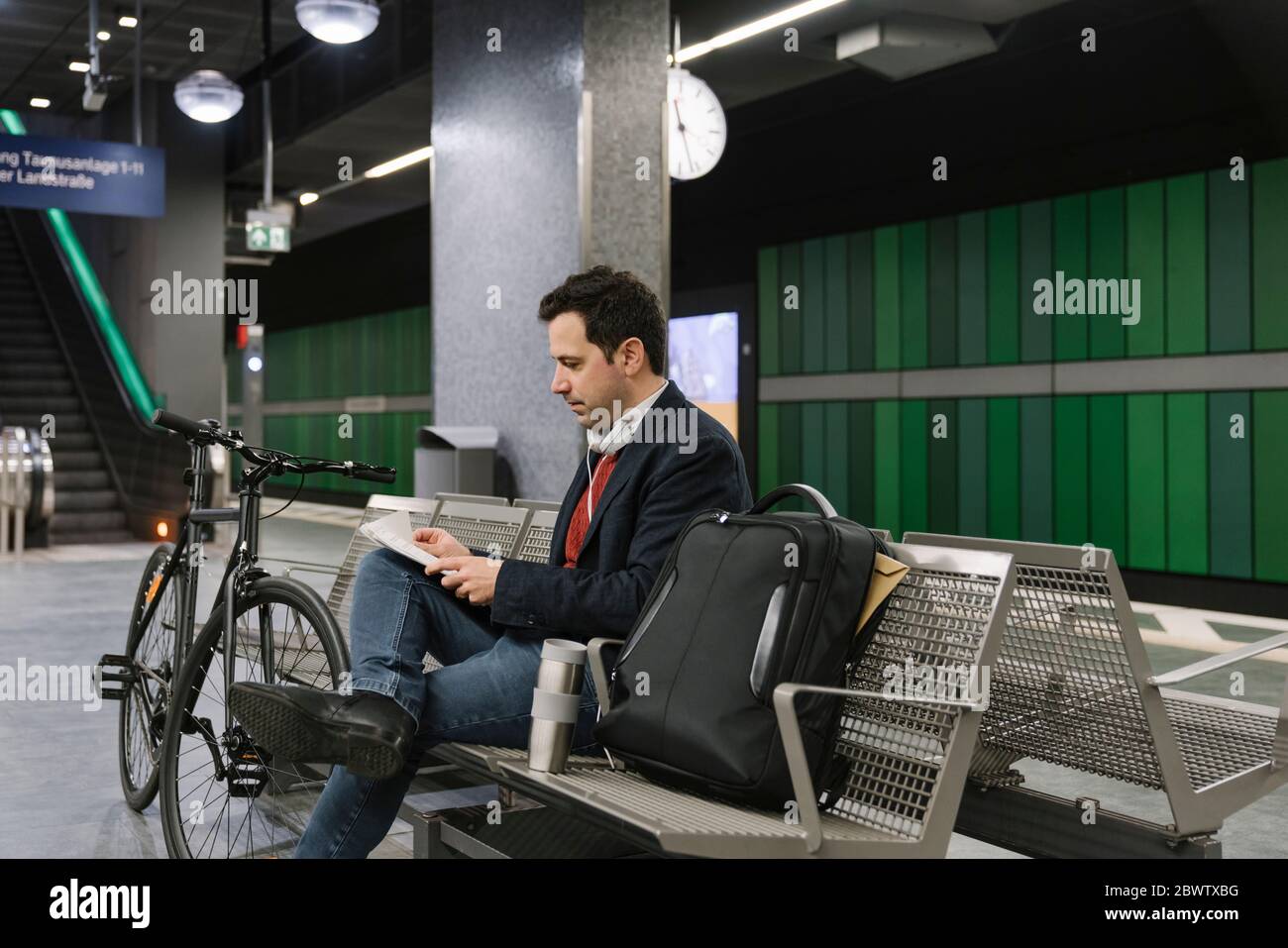 Geschäftsmann mit Fahrrad-Leseunterlagen, während er auf der Bank am U-Bahnhofsplatz sitzt, Frankfurt, Deutschland Stockfoto