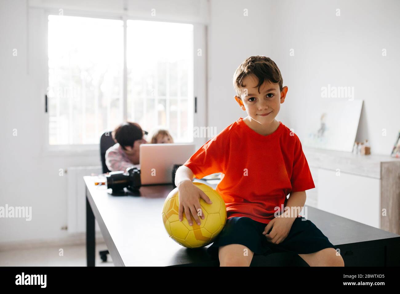 Mutter arbeitet zu Hause mit ihren Kindern, Sohn sitzt auf dem Tisch mit Fußball Stockfoto