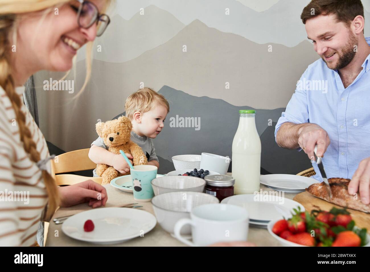 Kleiner Junge, der am Frühstückstisch sitzt und seinem Vater beim Brotschneiden zuschaut Stockfoto