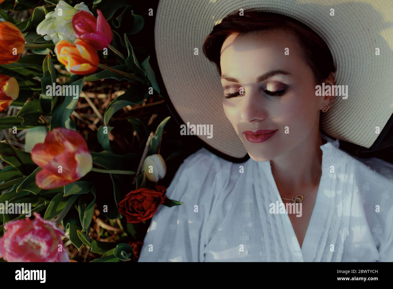 Portrait einer Frau mit geschlossenen Augen, die einen Hut auf dem Boden zwischen Tulpen trägt Stockfoto