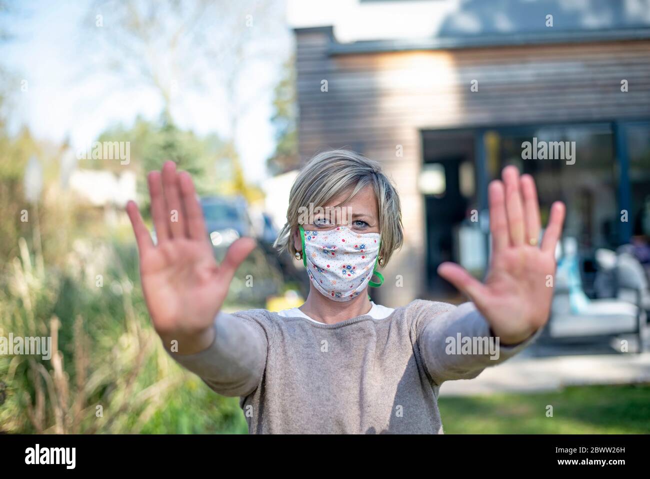 Portrait der reifen Frau trägt Schutzmaske Gesicht beim Stehen mit Stop-Geste gegen Haus während epidemische Lockdown Stockfoto