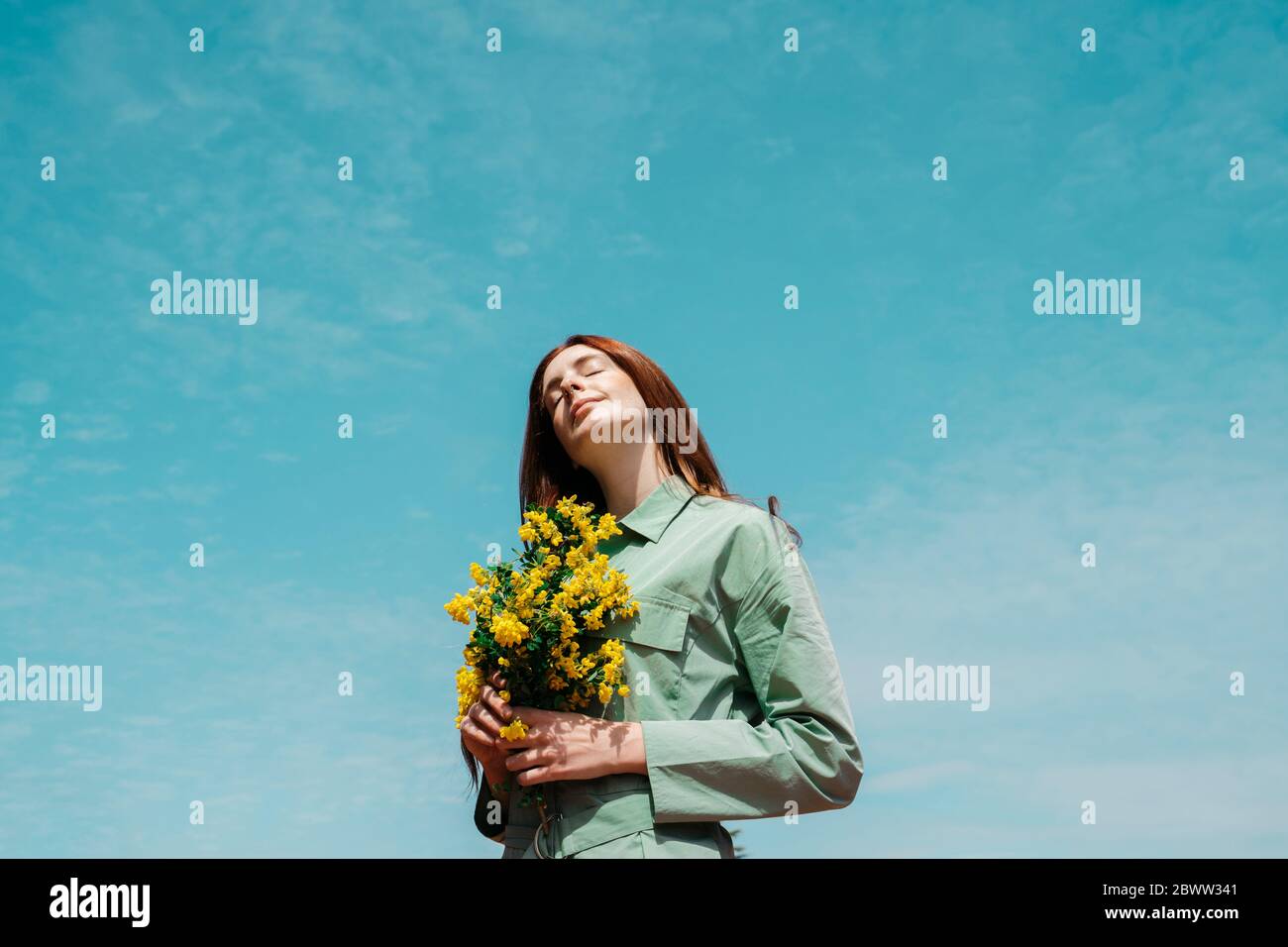 Porträt einer rothaarigen jungen Frau mit geschlossenen Augen, die gegen den Himmel steht und einen Strauß gelbter Blumen hält Stockfoto