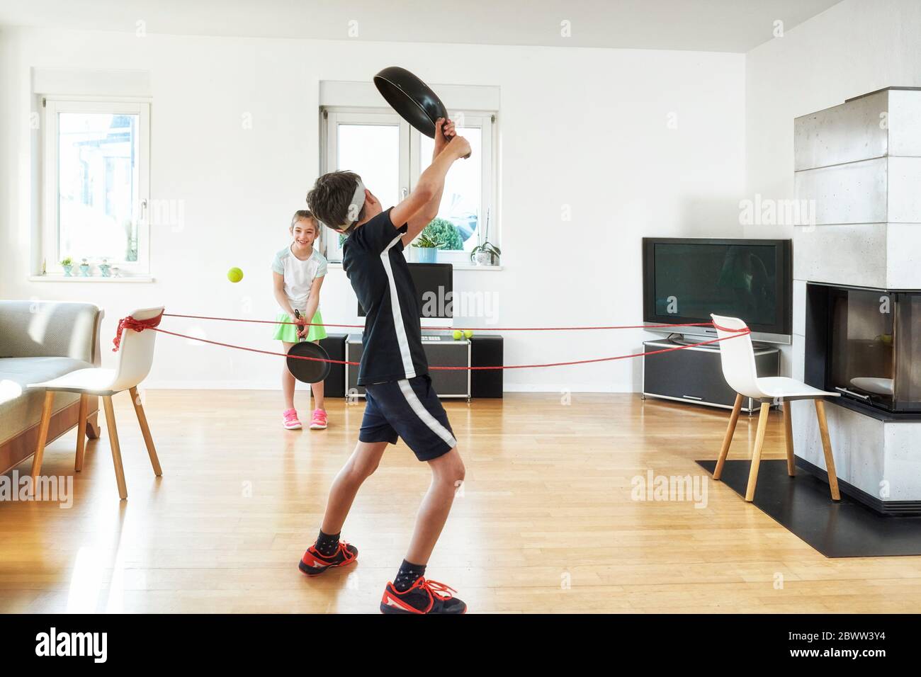 Geschwister spielen Tennis mit Bratpfannen im Wohnzimmer zu Hause während Quarantäne Stockfoto