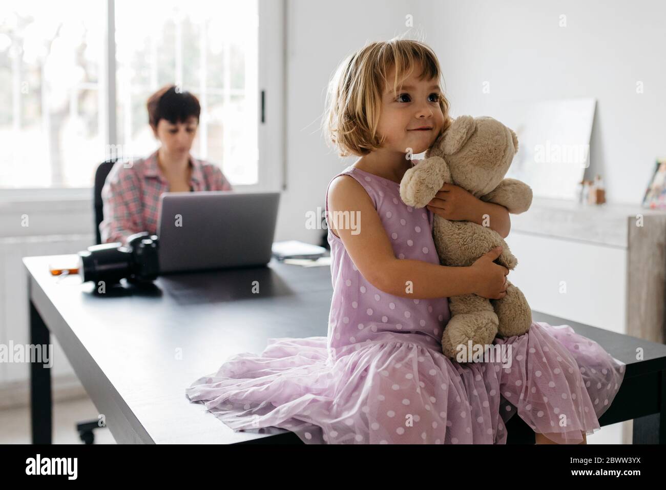 Mutter arbeitet zu Hause, Tochter sitzt mit Teddybär auf dem Tisch Stockfoto