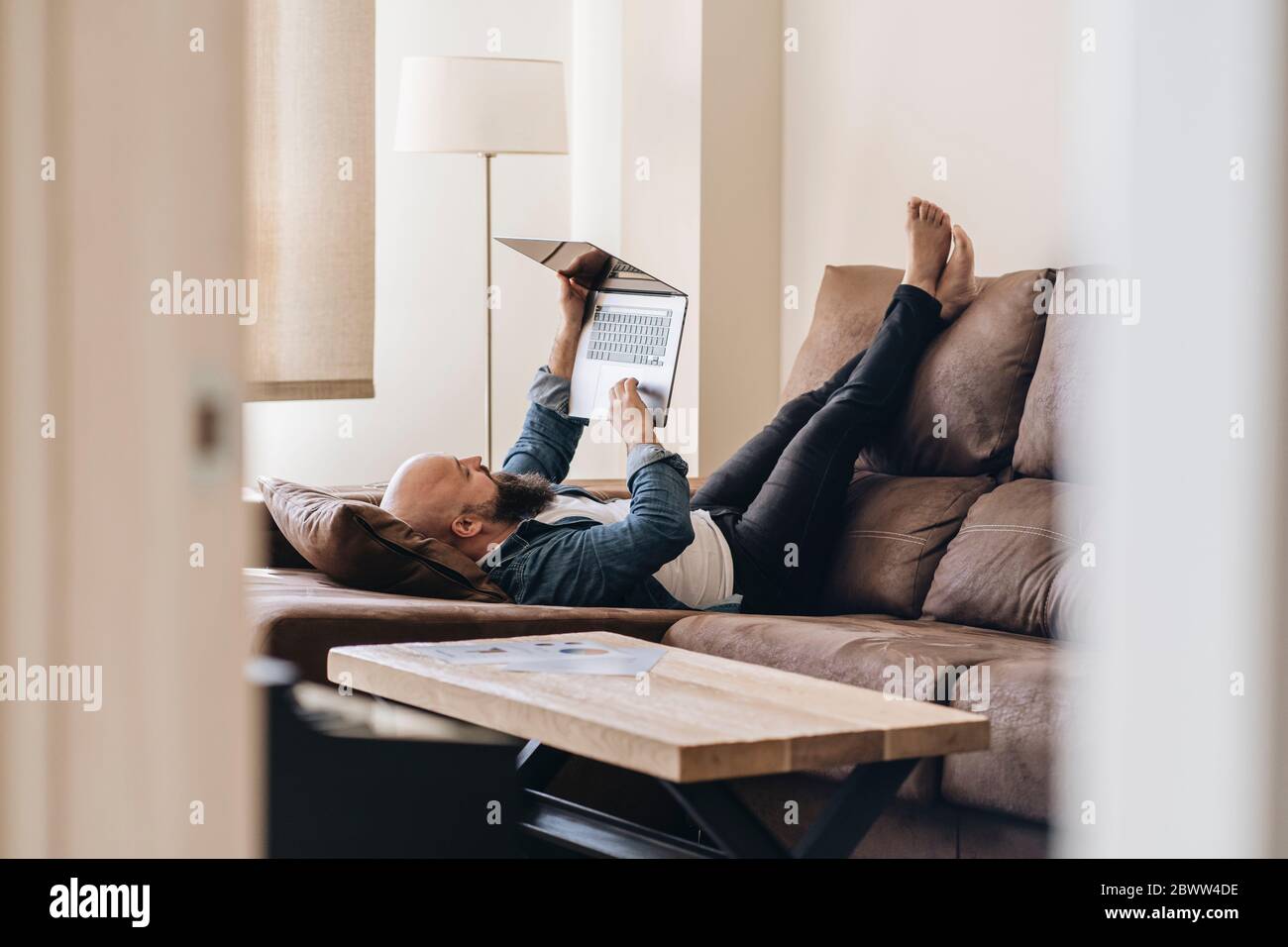 Gelangweilter Mann mit Laptop, während er im Wohnzimmer zu Hause auf dem Sofa lag Stockfoto